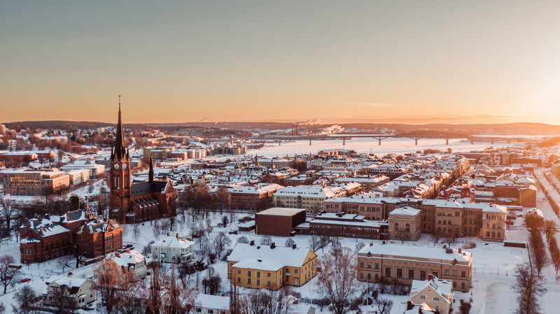 Drönarbild över Sundsvalls Stenstan och Sundsvallsbukten. Snö ligger över stadens gator och tak och solen är på väg upp över blå himmel.