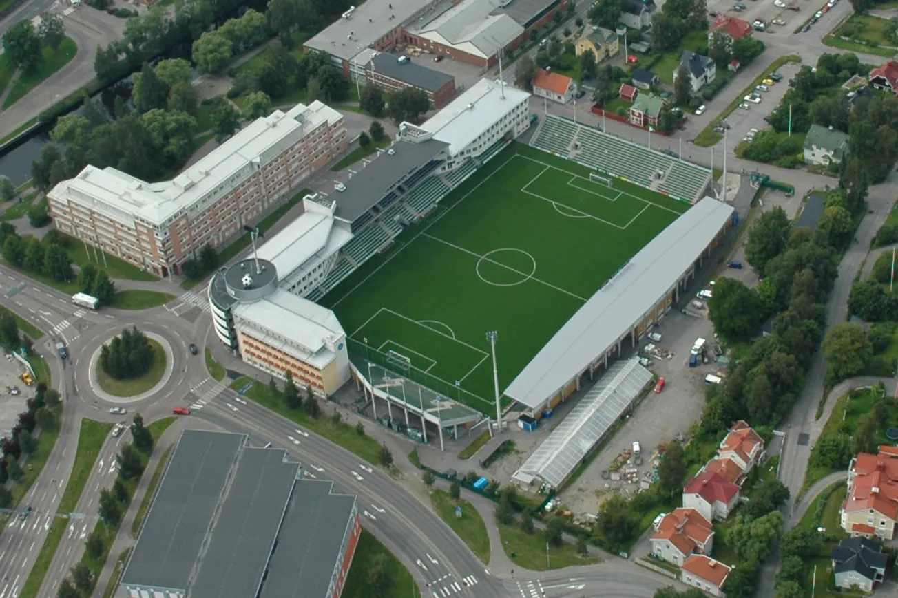 Flygfoto över en del av staden med en fotbollsarena i mitten med grön gräsmatta.