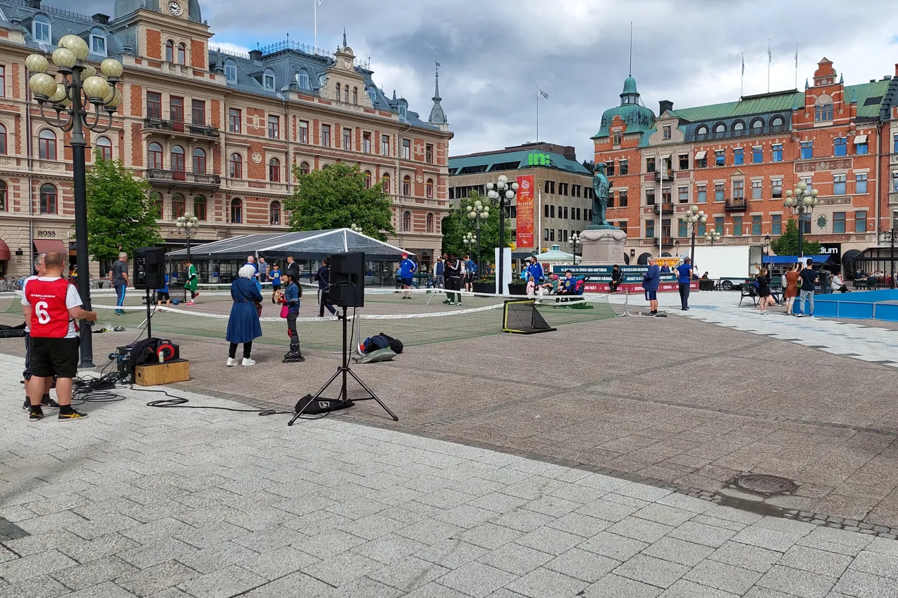 Stort torg med stenhus runt om, människor står och pratar i smågrupper.