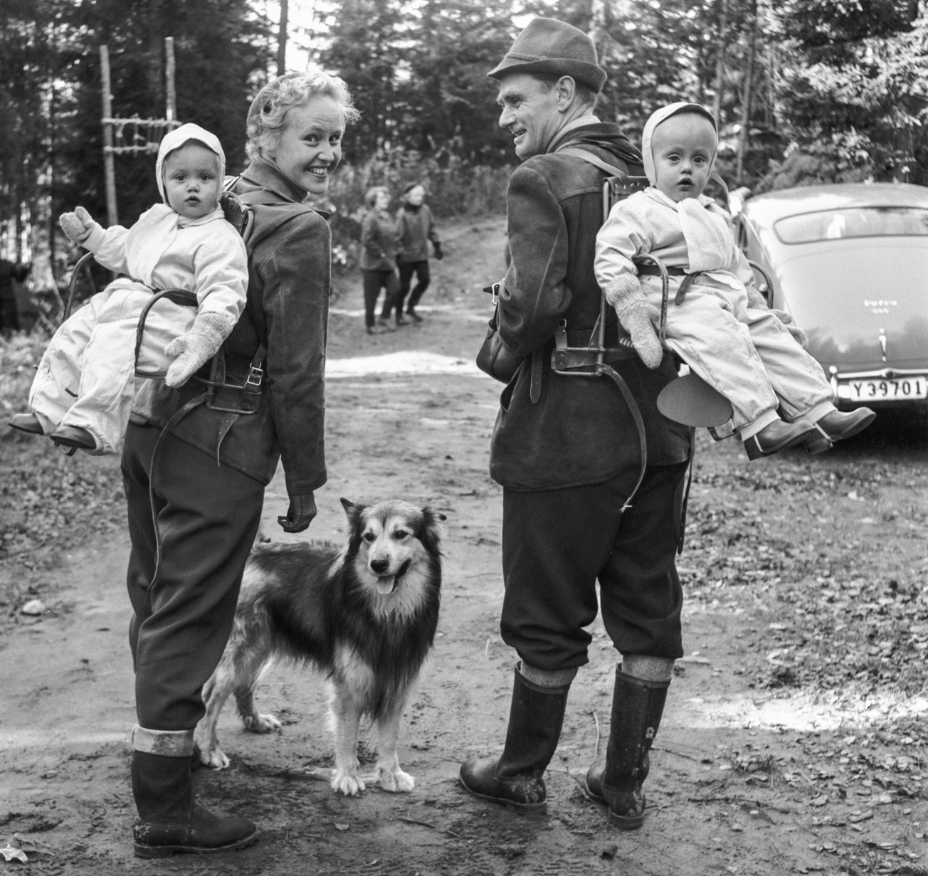 Två personer klädd för utflykt med hund och varsitt barn fastspänd på ryggen.