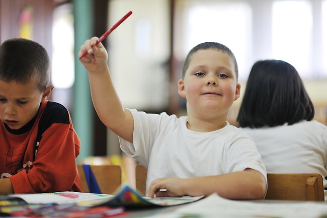 Ett barn sitter och arbetar med en skoluppgift och räcker upp handen. I handen håller hen en röd penna.