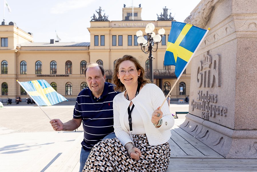 En man och en kvinna ler mot kameran och viftar med varsin svensk flagga som de håller i sina händer. I bakgrunden syns Stadshuset i Sundsvall.